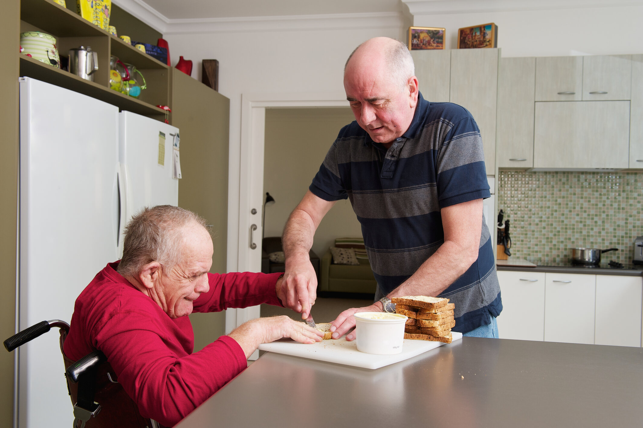 An older physically disabled man getting help making a sandwich in the kitchen.