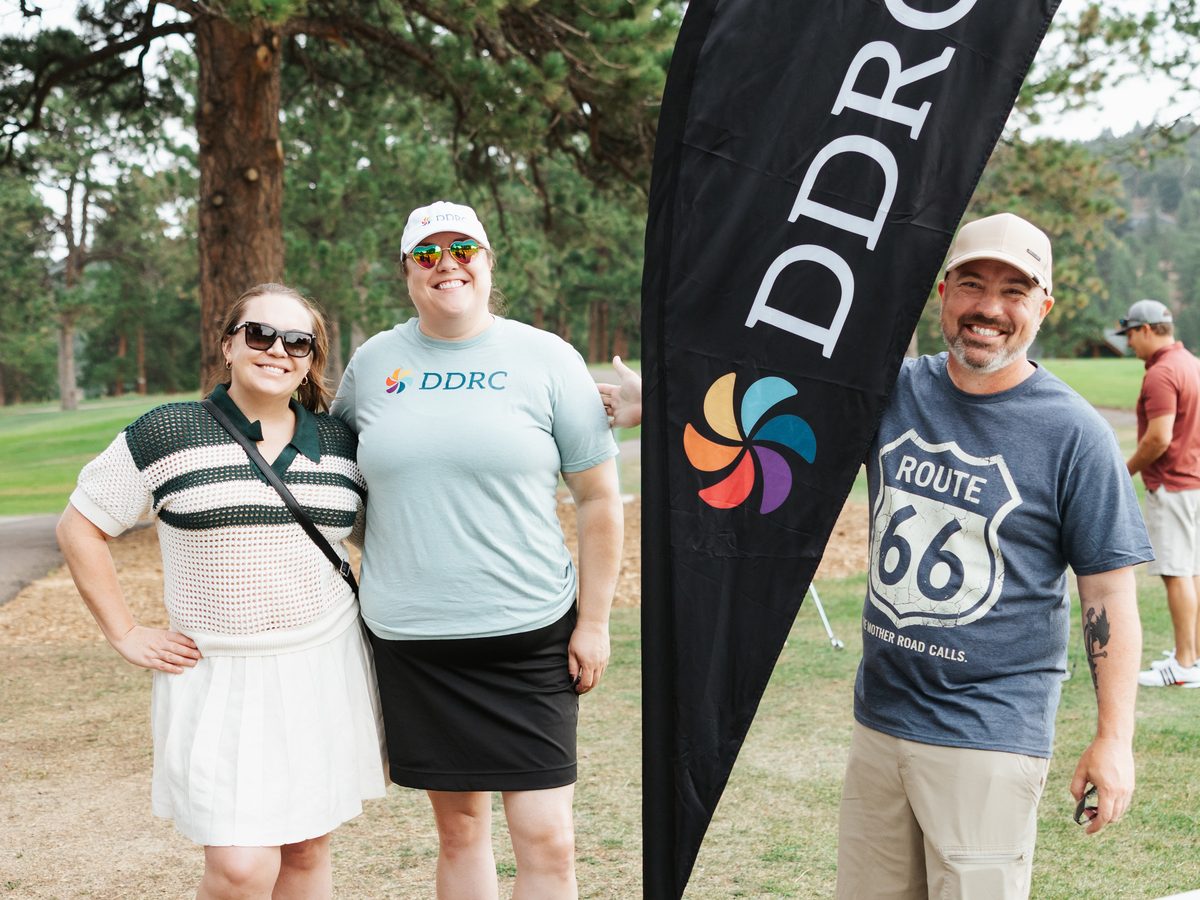 Three smiling guests pose beside a DDRC feather flag, one wearing a Route 66 T-shirt