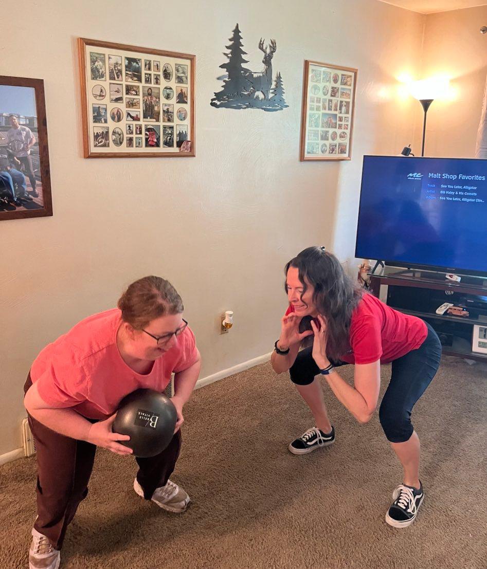 Amanda holds a weighted exercise ball at her knees in a squat position while Ruth squats alongside her, coaching the movement.
